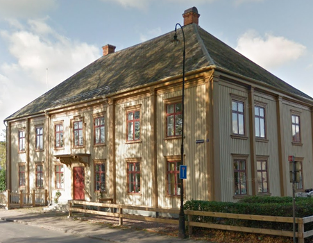 En stor träbyggnad i två våningar med ett brant tak och flera fönster, belägen i ett gathörn, med ett trästaket i förgrunden.