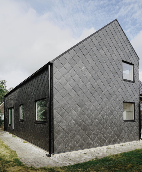 Ett modernt hus med ett brant sluttande tak och ytterväggar klädda med diamantmönstrade mörkgrå bältros, med flera rektangulära fönster och omgivet av en gångväg i sten.