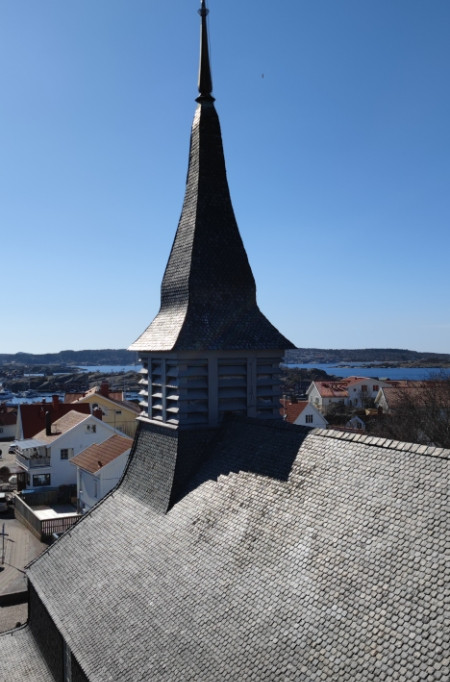 Ett högt kyrktorn med grå bältros reser sig över hustaken i en kuststad, med blått vatten och ett kuperat landskap synligt i bakgrunden under en klar himmel.