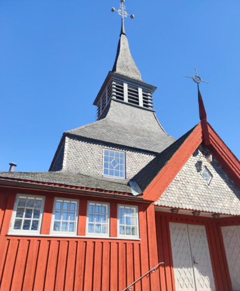 En röd träkyrka med vitkantade fönster och dörrar, med ett högt, grått shingled-torn och en väderflöjel på toppen, mot en klarblå himmel.
