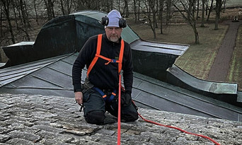 En person i säkerhetsutrustning, sele och hörlurar knäböjer på ett sluttande tak av metall och sten. Lövlösa träd och ett gräsbevuxet område syns i bakgrunden.
