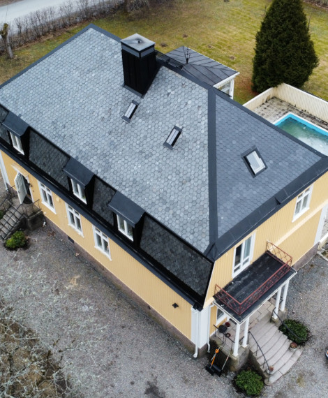 Flygfoto över ett tvåvåningshus i gult med ett mörkgrått hexagonalt mönstrat tak, flera takfönster, en liten balkong ovanför entrén och en inhägnad rektangulär swimmingpool på bakgården.
